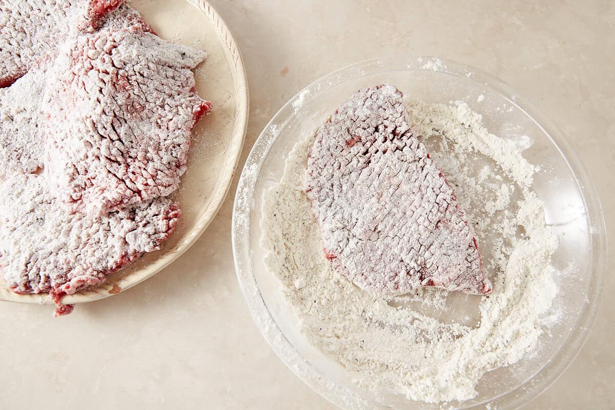 Raw pieces of meat coated in flour sit on a plate, with one piece being dredged in a dish of flour on a light-colored countertop.