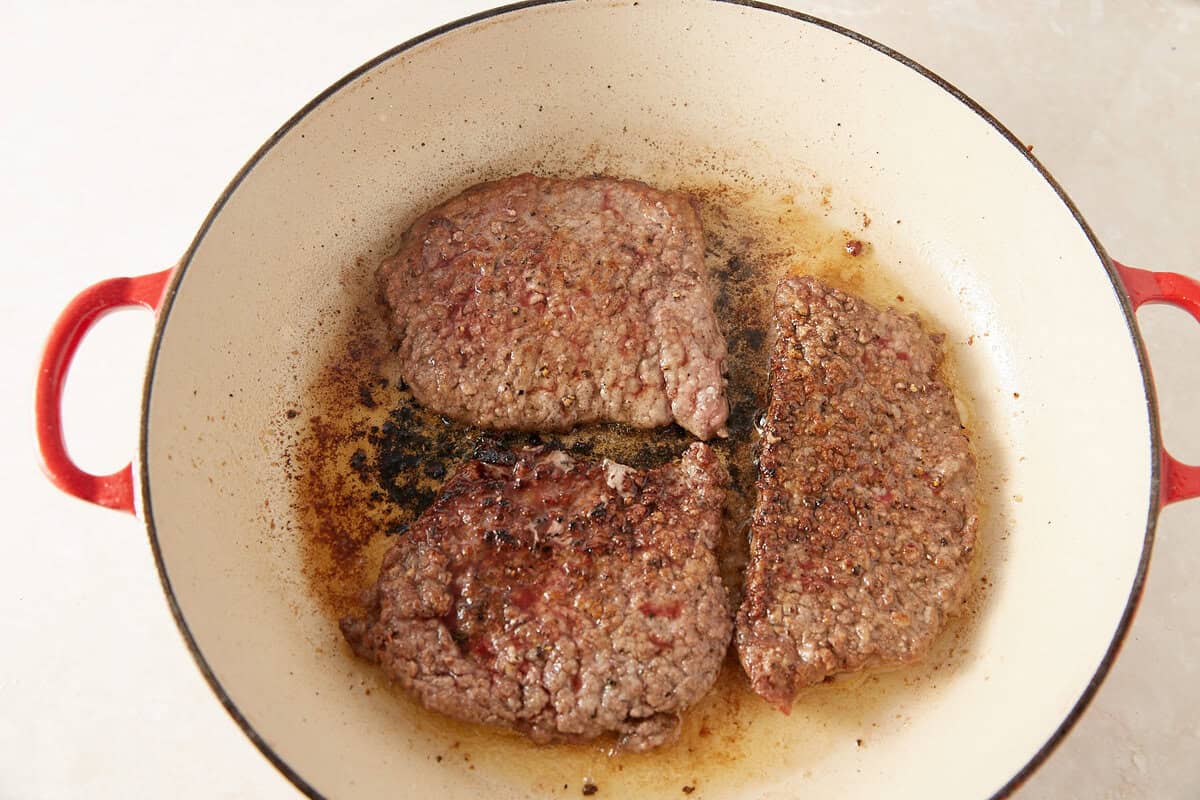 Three browned ground beef patties are cooking in a white, round skillet with red handles. The patties are sizzling in rendered fat, and the pan shows some browning and crispy residue.