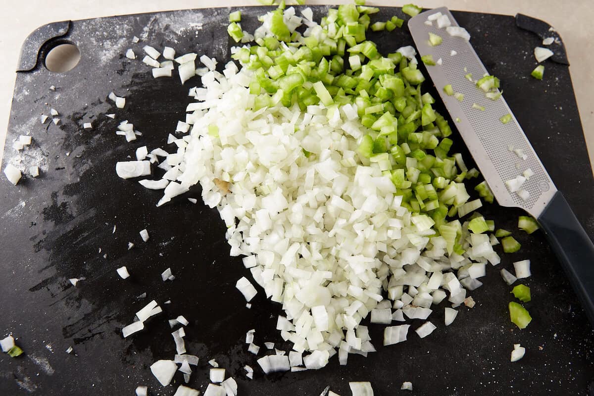 Chopped white onions and green celery on a black cutting board with a knife beside them.