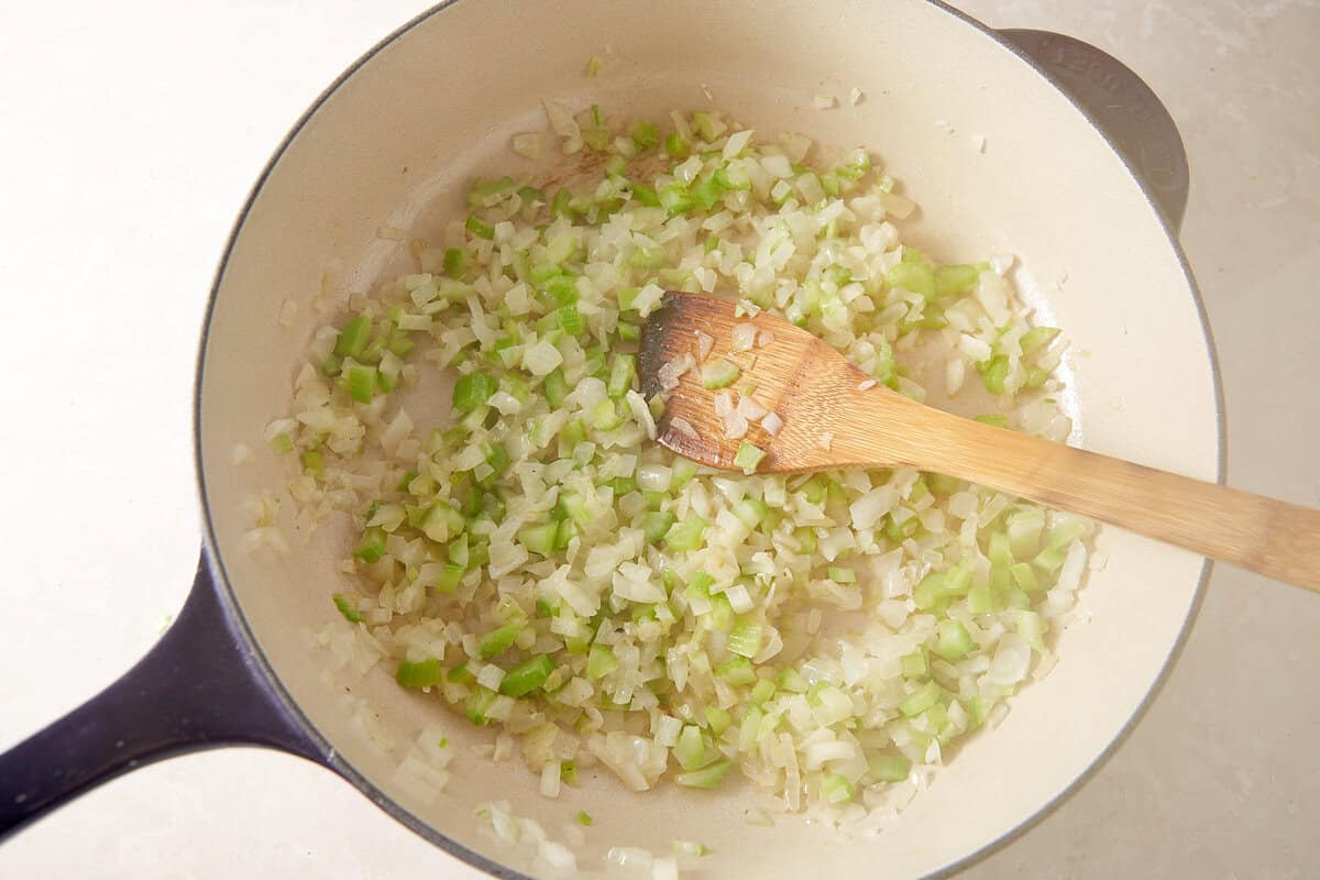 Chopped onions and celery are being sautéed in a large light-colored pot, stirred with a wooden spoon.