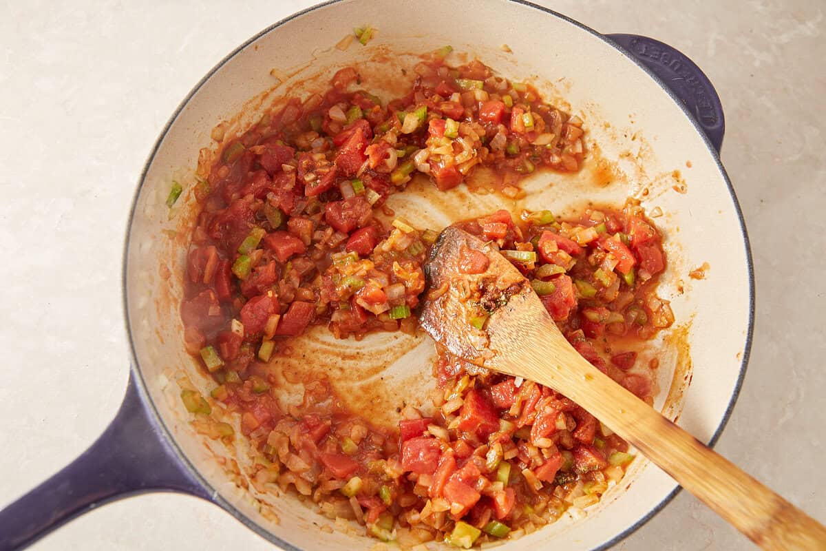 A blue pot sits on a light countertop, filled with a mixture of sautéed diced tomatoes, onions, and green bell peppers. A wooden spoon rests inside, stirring the colorful, partially cooked vegetables.