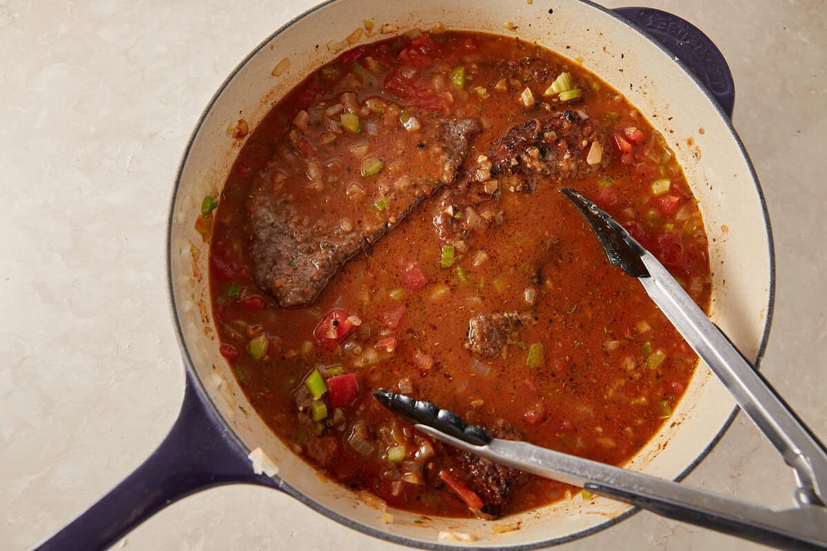 A pot of braised meat in a tomato-based sauce with chopped vegetables, accompanied by metal tongs resting on the edge of the pot.