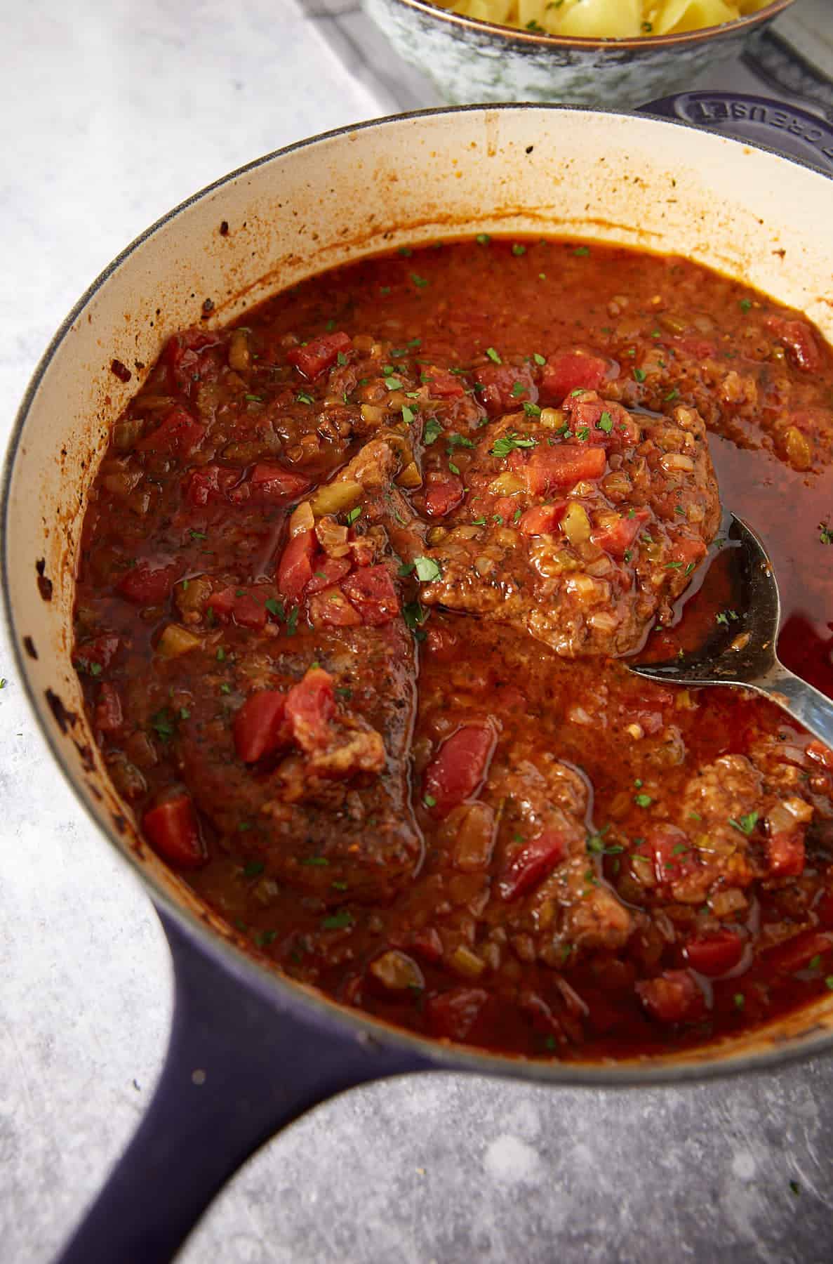 A cast iron pan filled with beef steaks simmered in a chunky tomato-based sauce with chopped onions and herbs, placed on a light surface. A spoon rests in the sauce next to the steaks.