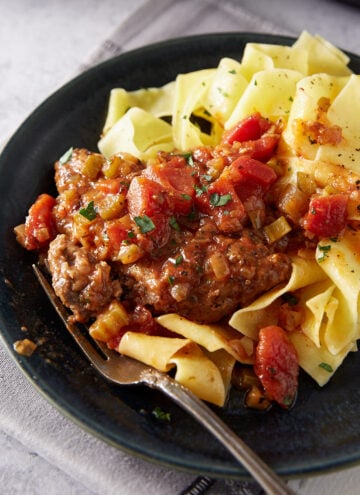 A black plate with wide egg noodles topped with chopped herbs and served alongside a hamburger steak in tomato and vegetable sauce, with a fork resting on the plate.
