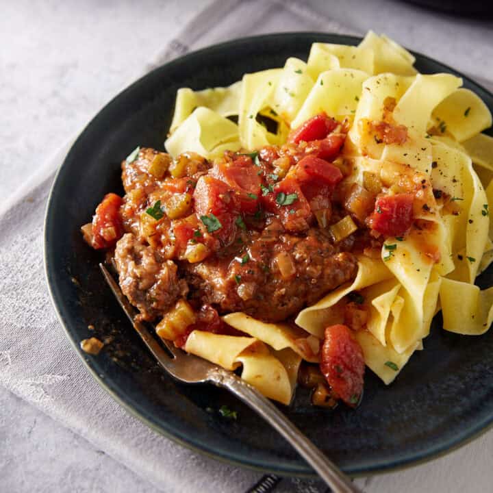 A black plate with wide egg noodles topped with chopped herbs and served alongside a hamburger steak in tomato and vegetable sauce, with a fork resting on the plate.
