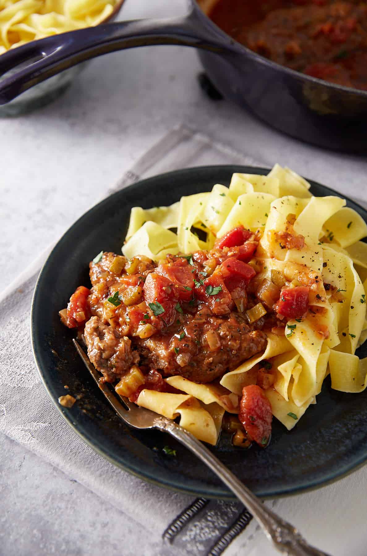 A plate of wide pasta noodles topped with a braised meat dish, covered in a chunky tomato and vegetable sauce, served with a fork on a dark plate. A pot of more sauce is visible in the background.
