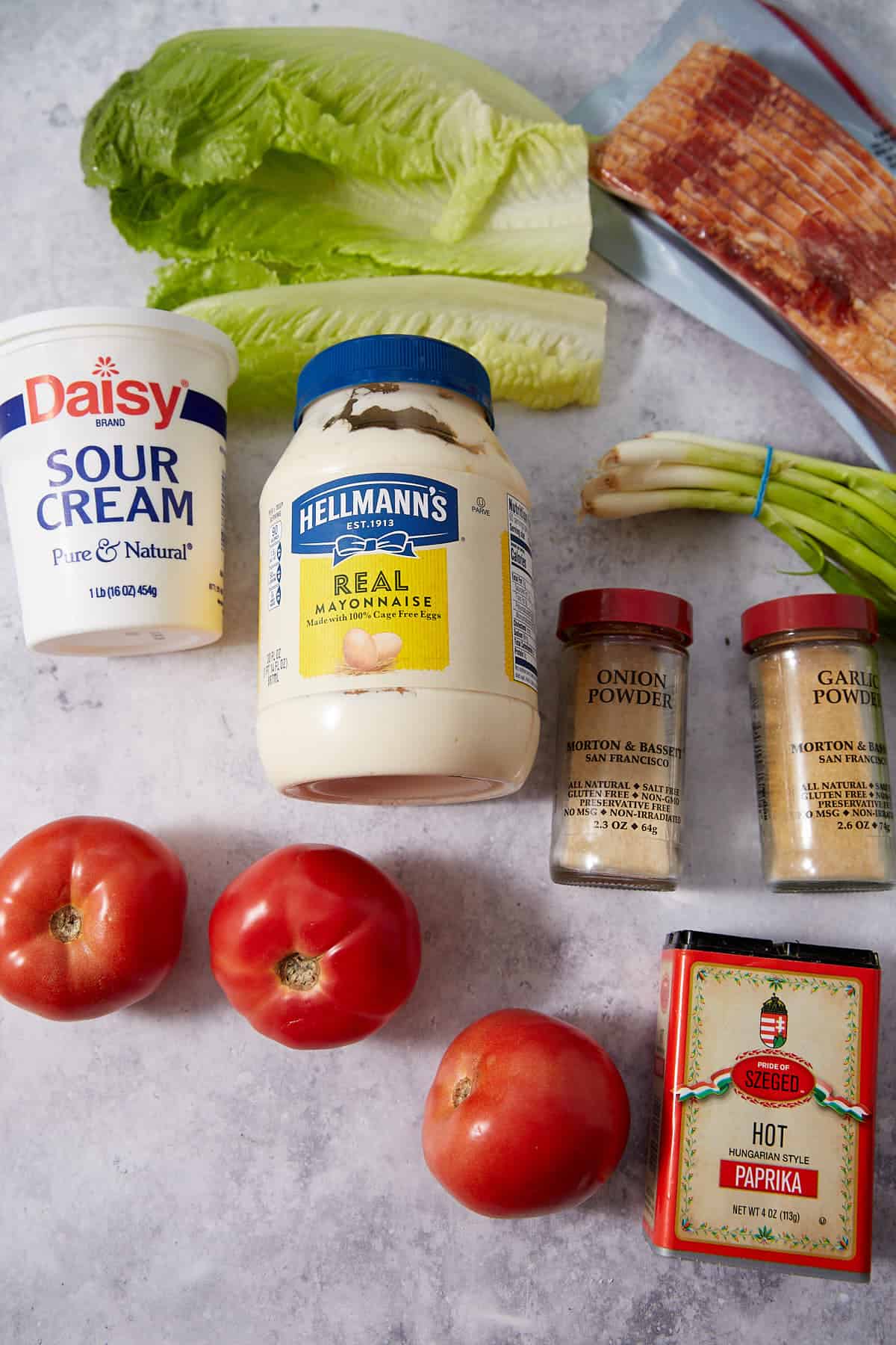 Various ingredients on a countertop, including sour cream, mayonnaise, paprika, onion and garlic powder, bacon, romaine lettuce, green onions, and three tomatoes.