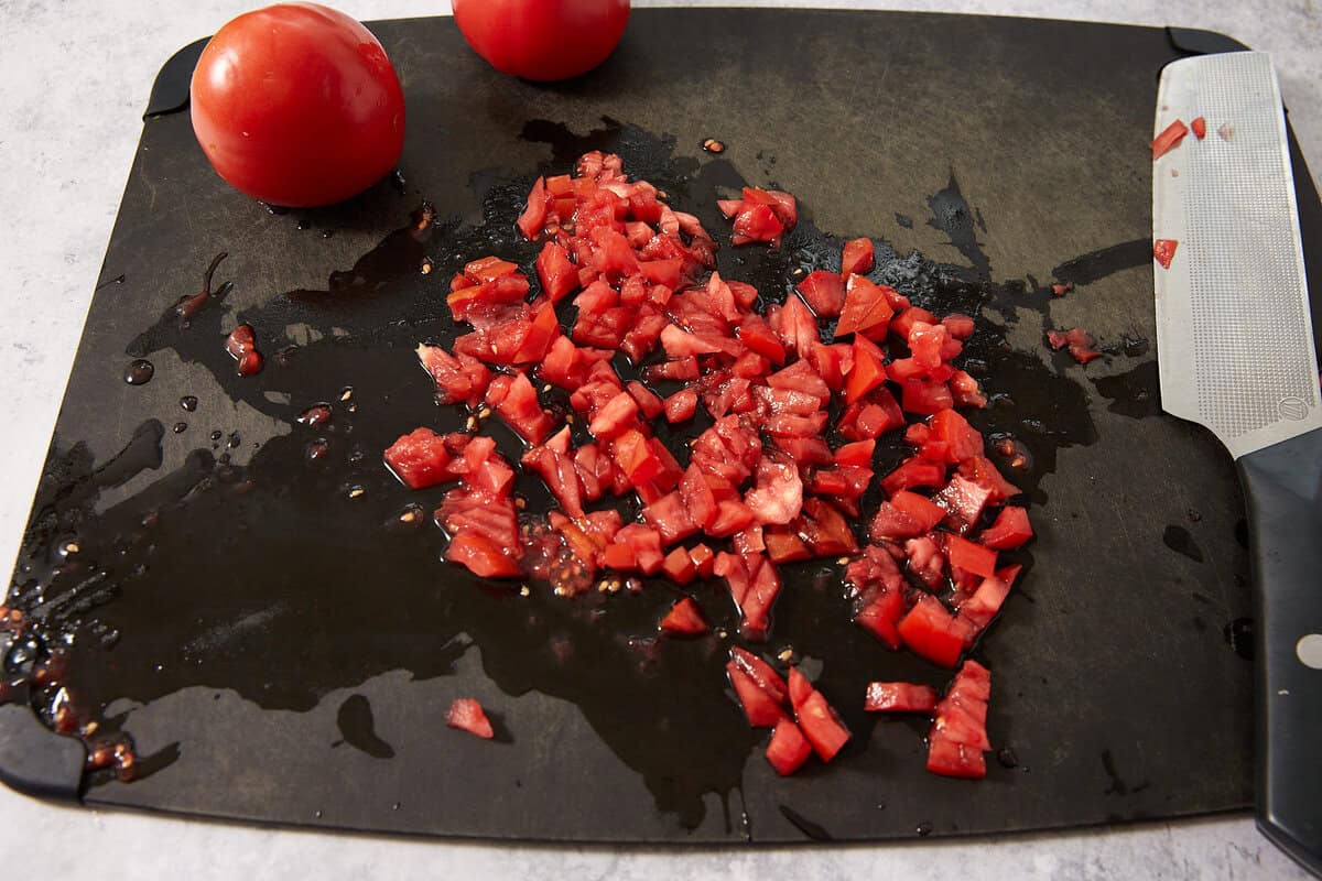 Diced tomatoes on a black cutting board with two whole tomatoes and a knife nearby; tomato juice is visible on the board.