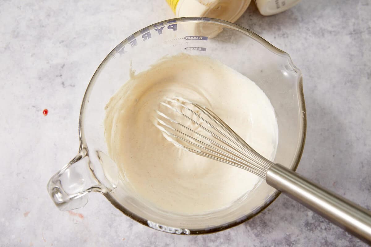 A metal whisk sits in a glass measuring cup filled with creamy white sauce, placed on a light gray countertop.