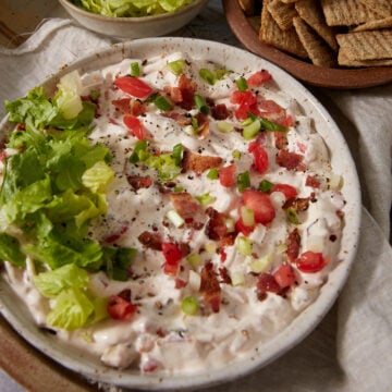 Bowl of creamy dip topped with chopped lettuce, tomatoes, green onions, and bacon bits. Crackers and extra shredded lettuce are placed nearby on the table.
