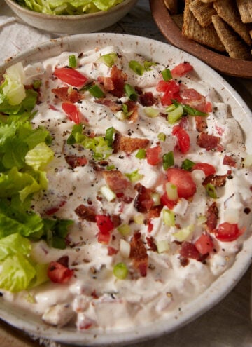 Bowl of creamy dip topped with chopped lettuce, tomatoes, green onions, and bacon bits. Crackers and extra shredded lettuce are placed nearby on the table.