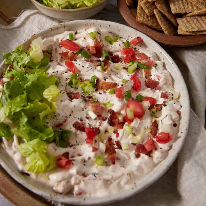 Bowl of creamy dip topped with chopped lettuce, tomatoes, green onions, and bacon bits. Crackers and extra shredded lettuce are placed nearby on the table.