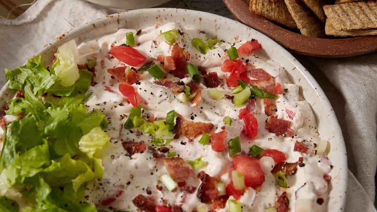 A plate of creamy dip topped with lettuce, tomatoes, bacon, and green onions