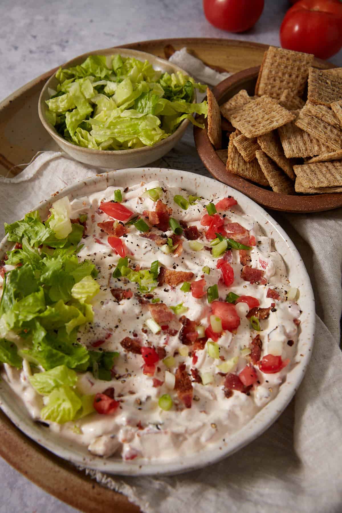 A plate of creamy dip topped with lettuce, tomatoes, bacon, and green onions, served with a bowl of shredded lettuce and a plate of square crackers on a beige tray.