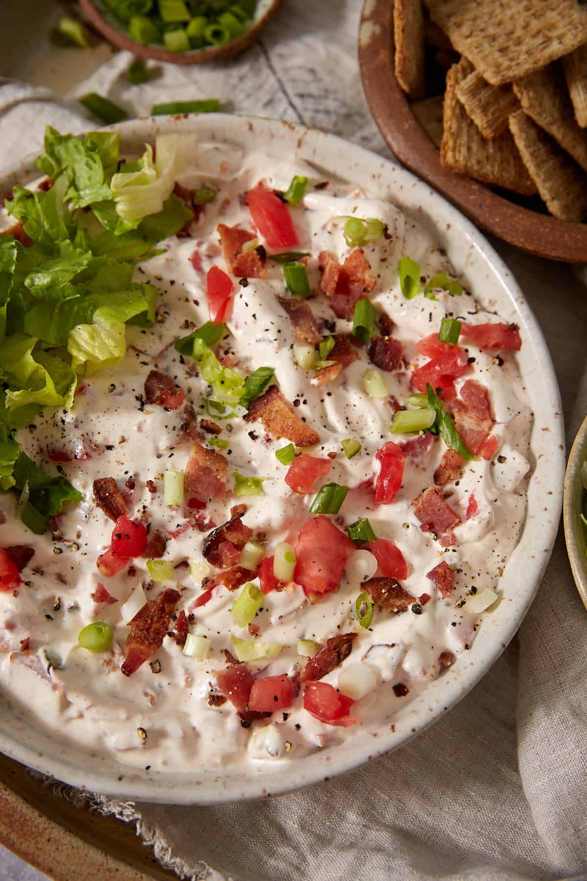 A bowl of creamy dip topped with chopped lettuce, tomatoes, bacon pieces, and green onions, served with a side of whole grain crackers.