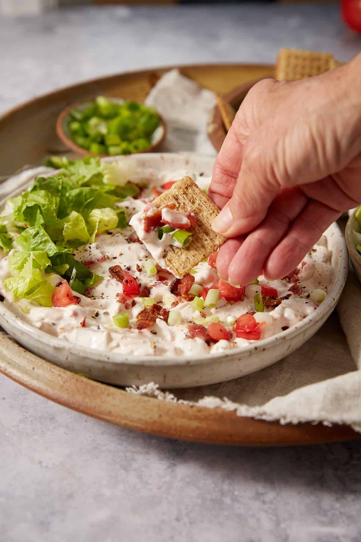 A hand dips a cracker into a creamy dip topped with lettuce, diced tomatoes, and green onions. More crackers and chopped green onions are in small bowls in the background.