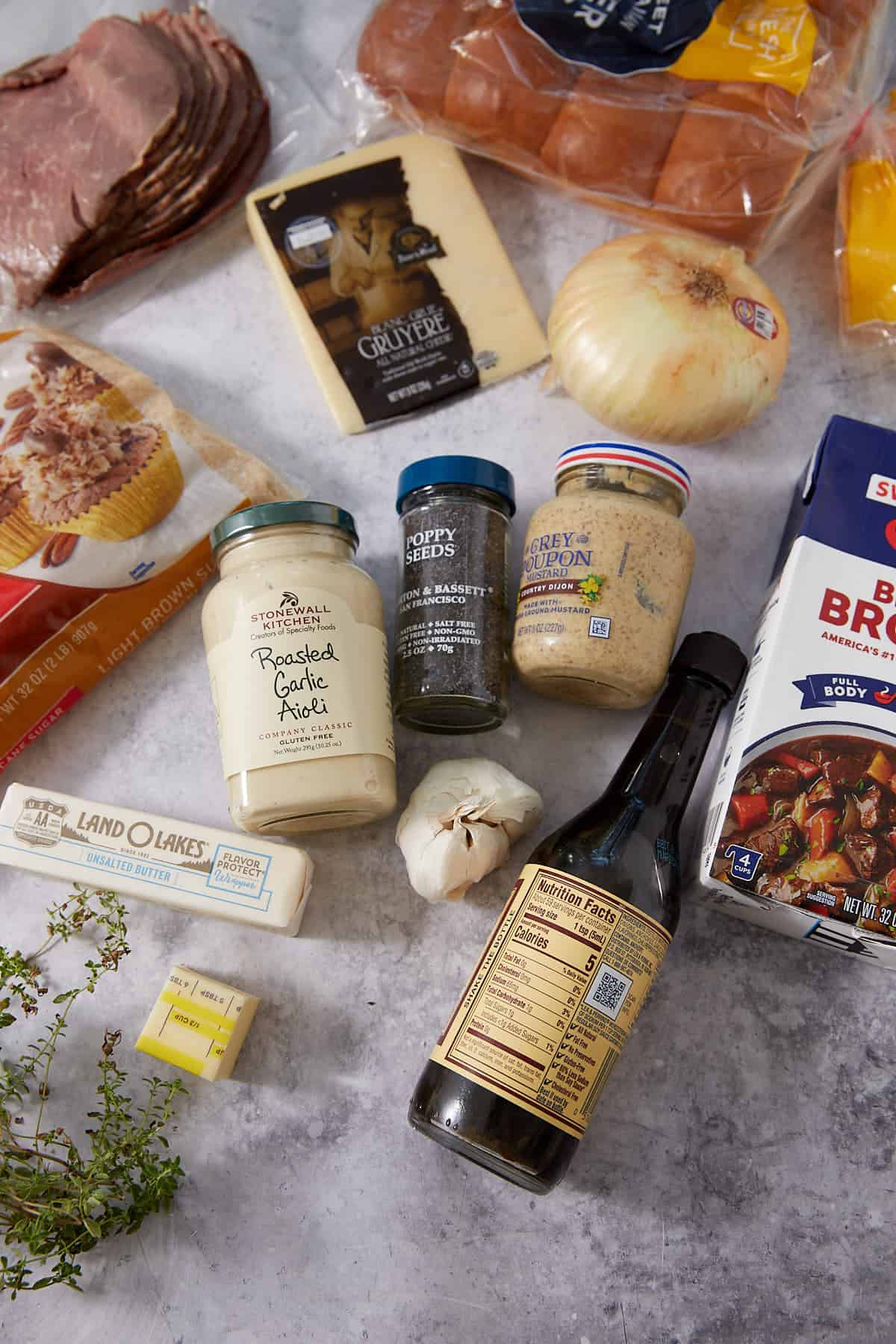 Various sandwich ingredients on a countertop, perfect for preparing French dip sliders— including bread, sliced roast beef, cheese, onion, butter, roasted garlic aioli, poppy seeds, Dijon mustard, Worcestershire sauce, garlic, and fresh herbs.