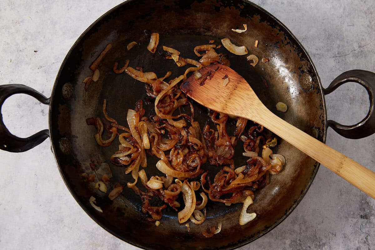 Sliced onions being caramelized in a dark skillet, with a wooden spoon resting on top of the onions—perfectly prepped for savory French dip sliders.