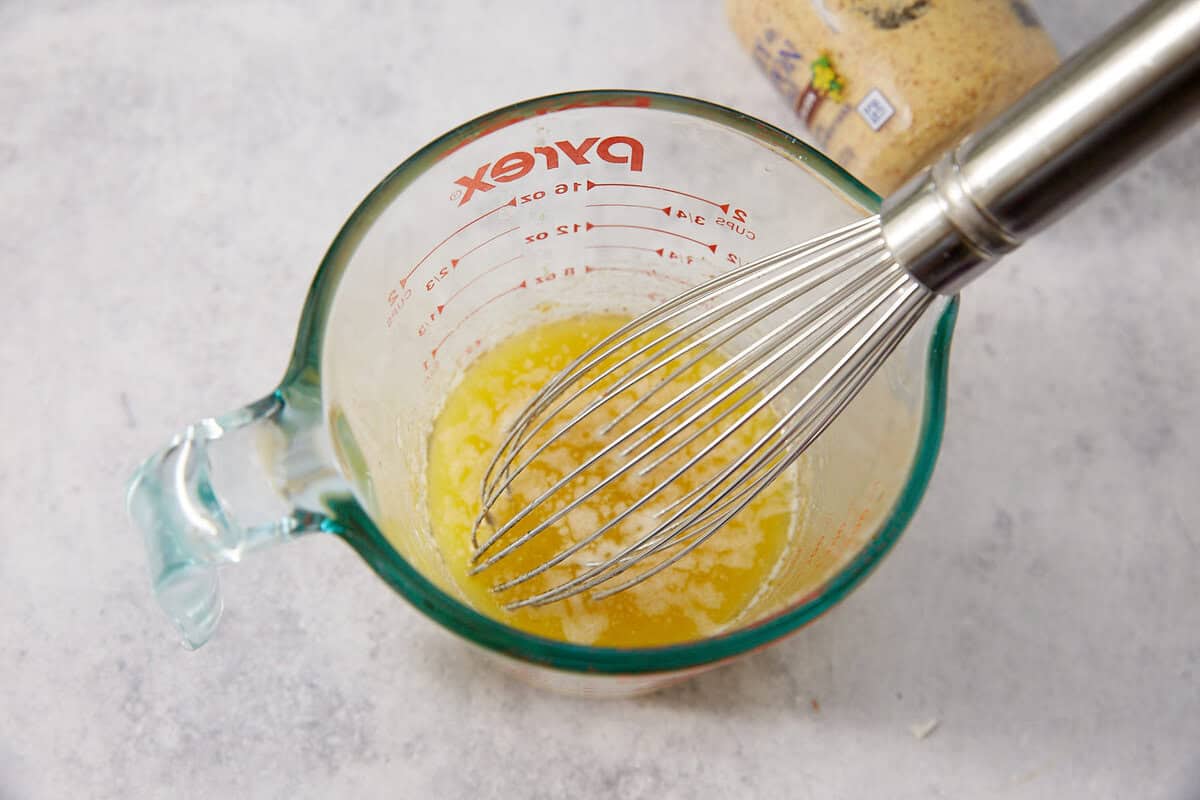 A metal whisk is mixing melted butter in a glass Pyrex measuring cup, placed on a light countertop—perfect for preparing French dip sliders. A container of breadcrumbs is partially visible in the background.