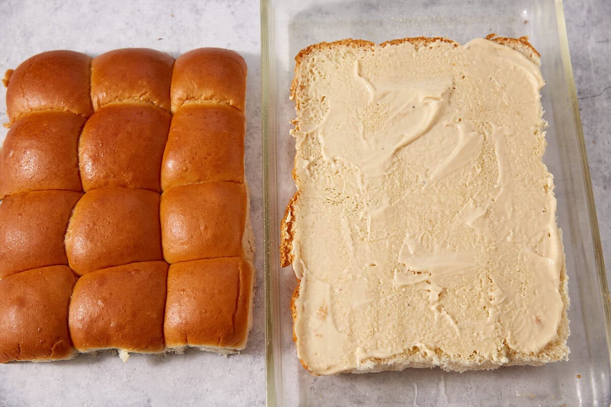 A set of connected slider buns is next to a glass dish holding the bottom half of the buns, which are spread with a light-colored sauce or mayonnaise, ready to be assembled into delicious French dip sliders.