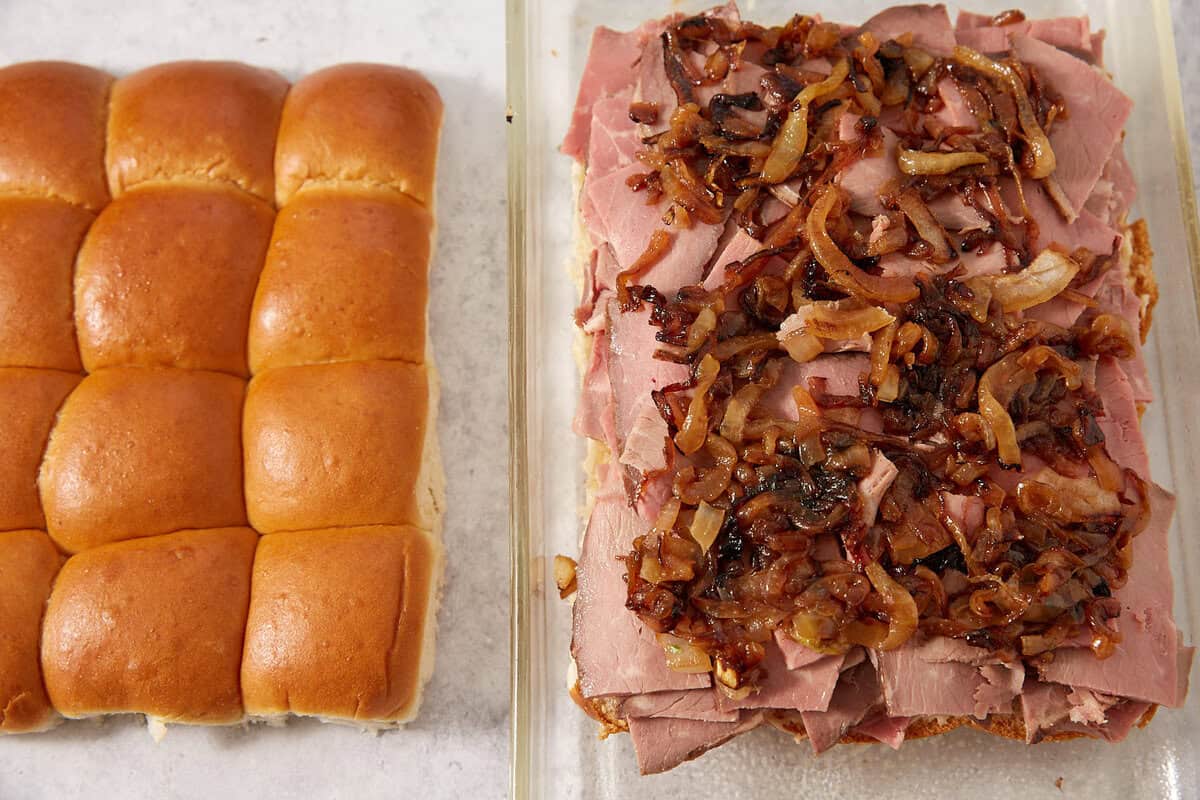 A tray of slider buns sits next to an open sandwich with layers of sliced roast beef and caramelized onions on top, ready to be assembled into savory French dip sliders.