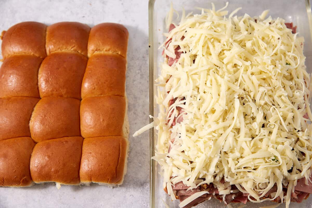 On the left, a tray of intact slider buns; on the right, an open French dip sliders dish filled with layers of shredded cheese, deli meat, and the bun bottoms, ready for baking.