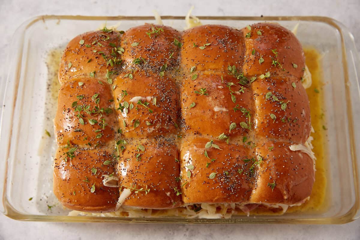 A glass baking dish holds a row of golden baked French dip sliders topped with poppy seeds and fresh herbs, with melted cheese visible along the edges.