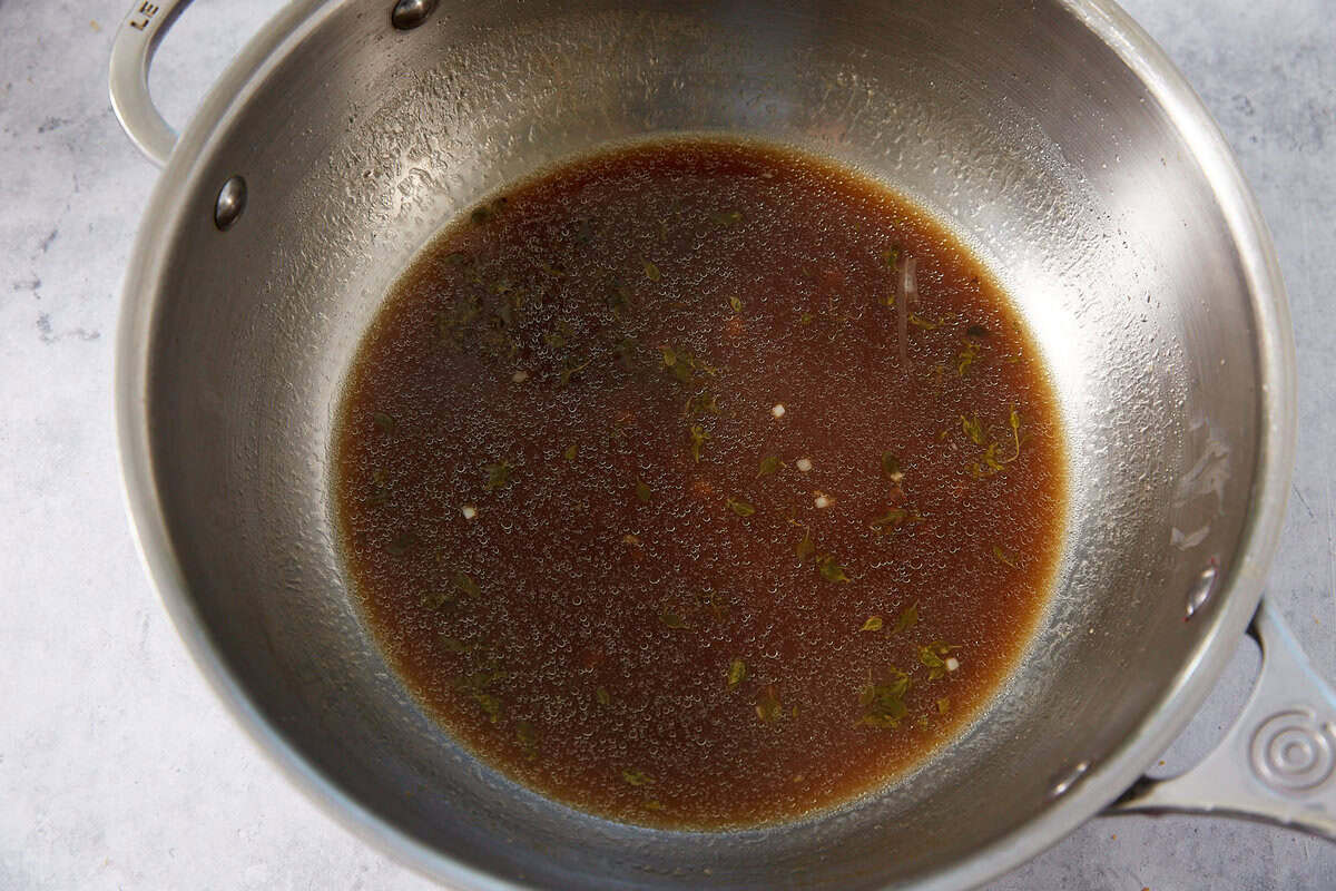 A metal pot with a small amount of brown broth or sauce, reminiscent of the savory au jus served with French dip sliders, containing visible herbs and small bits, sits on a light gray surface.