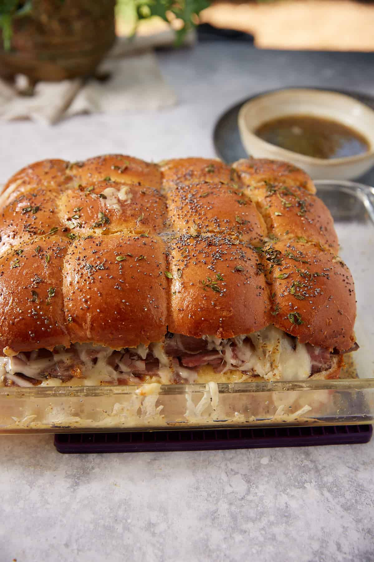 A glass dish holds freshly baked French dip sliders with golden buns, melted cheese, and visible meat layers, all topped with herbs and poppy seeds. A bowl of brown dipping sauce sits in the background.
