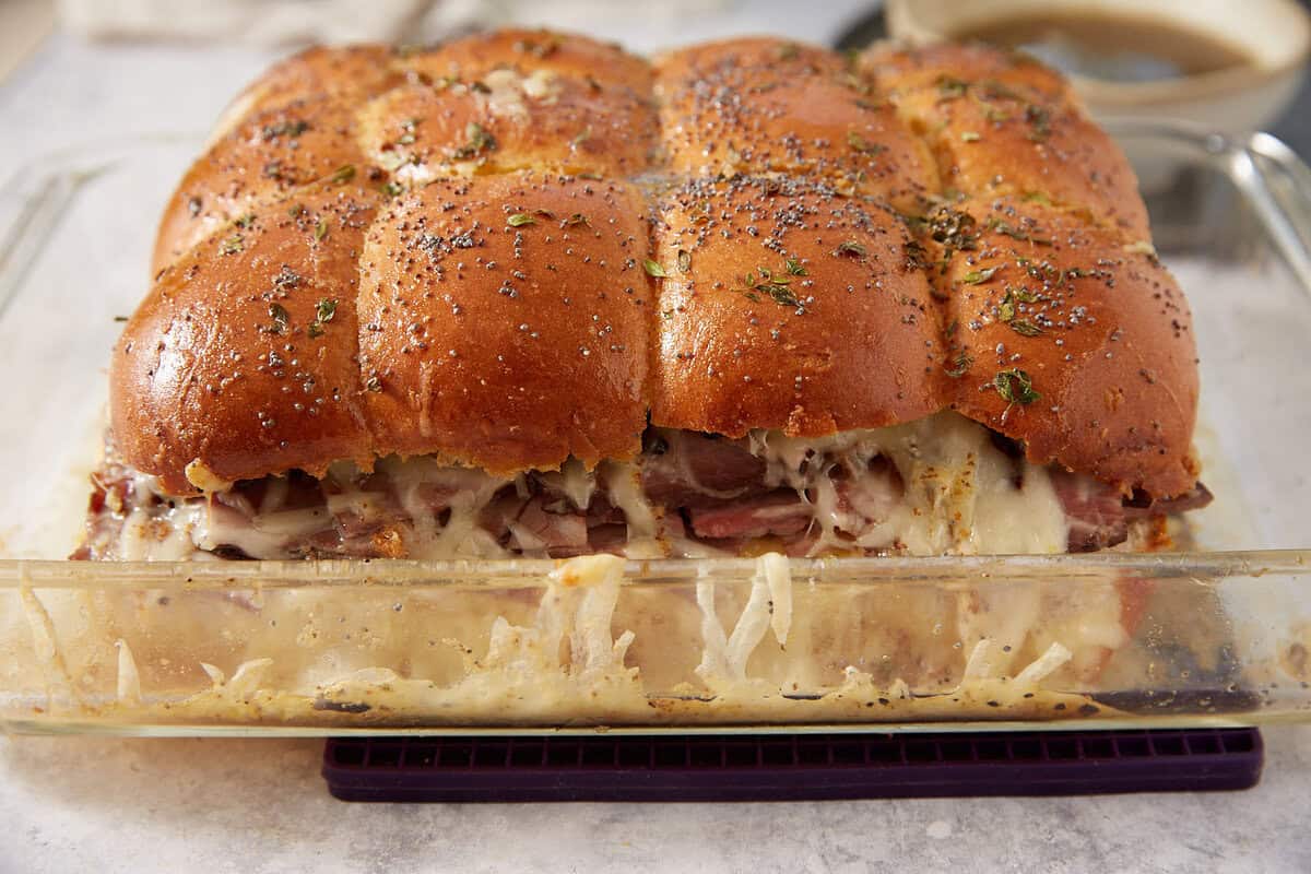 A glass baking dish filled with baked French dip sliders made with golden buns, melted cheese, and layers of meat, topped with herbs and poppy seeds.