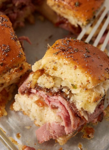 A close-up of French dip sliders with poppy seed topping, filled with layers of sliced meat and melted cheese, resting on a baking dish with a metal spatula underneath.