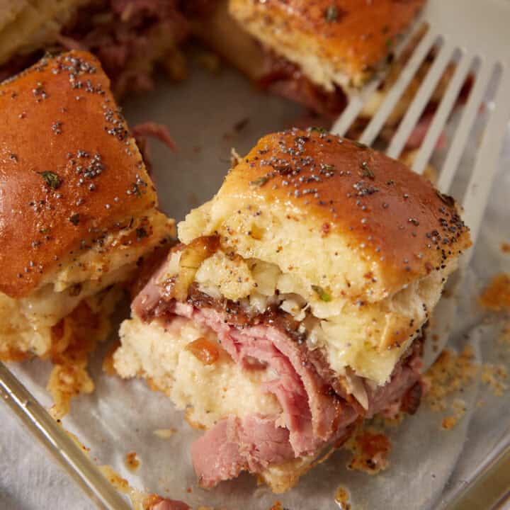 A close-up of French dip sliders with poppy seed topping, filled with layers of sliced meat and melted cheese, resting on a baking dish with a metal spatula underneath.