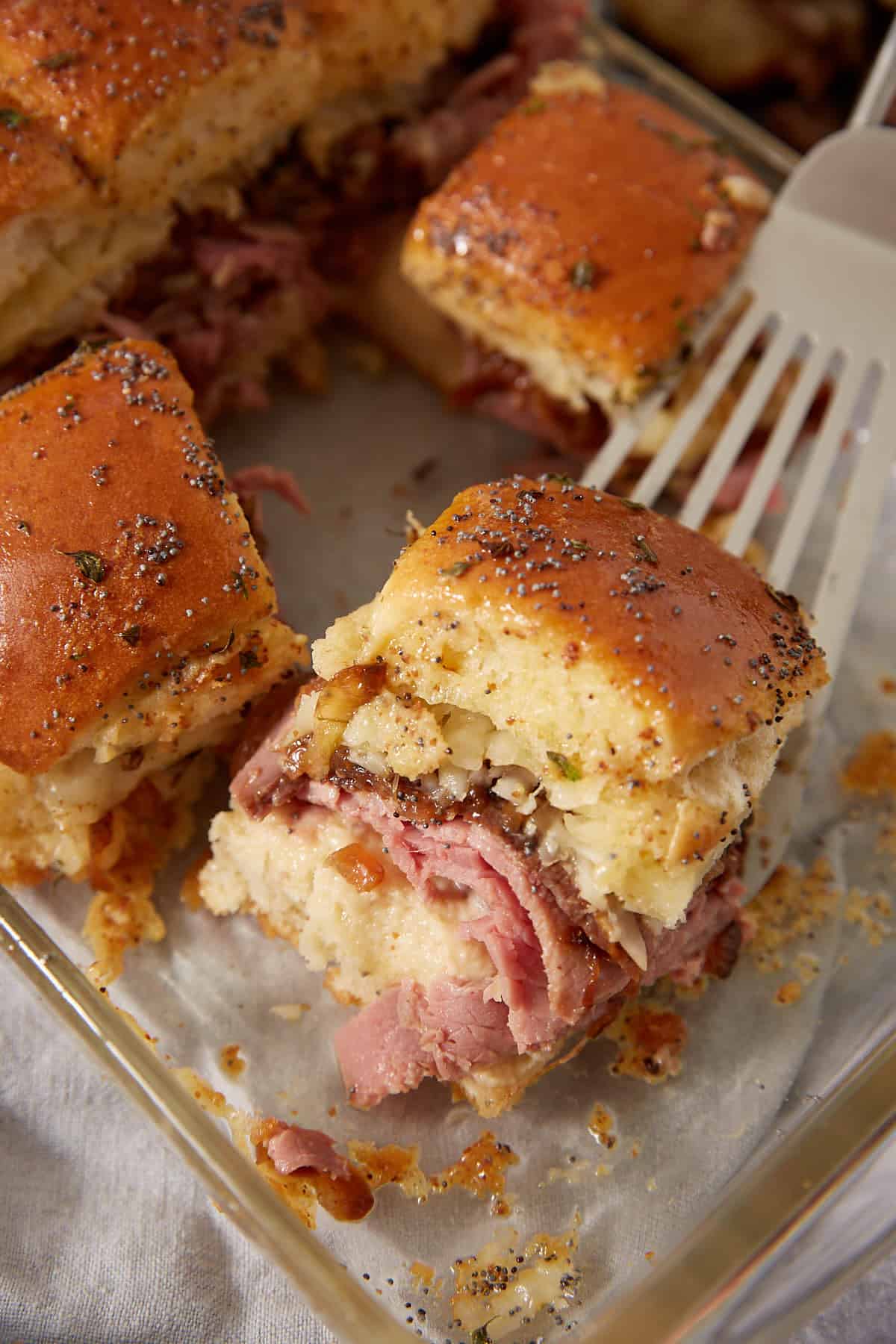 A close-up of a glass baking dish showcases savory French dip sliders filled with sliced meat, melted cheese, and topped with poppy seeds. A spatula is lifting one slider out from the dish.