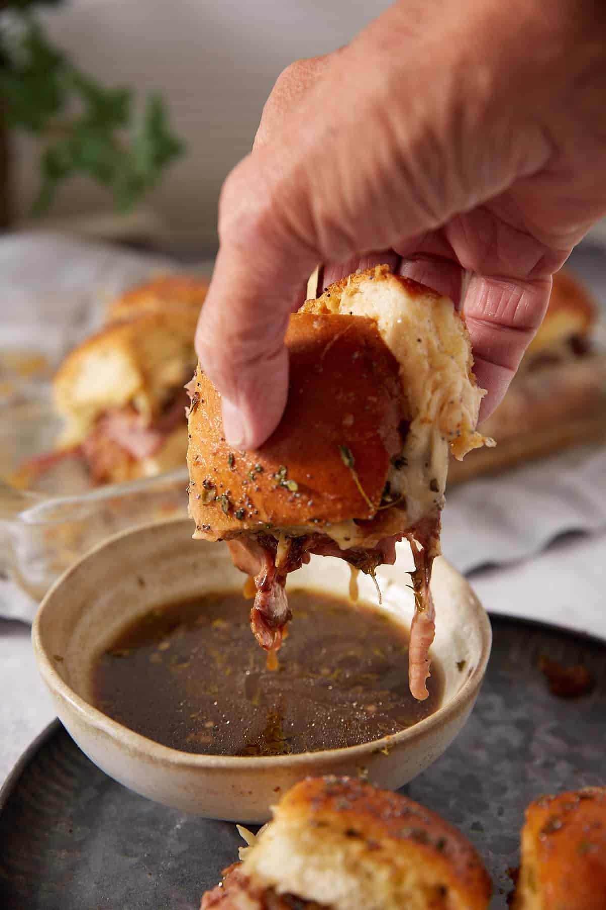 A hand dips a cheesy French dip slider sandwich into a bowl of au jus sauce, with more sliders and sauce visible in the background.