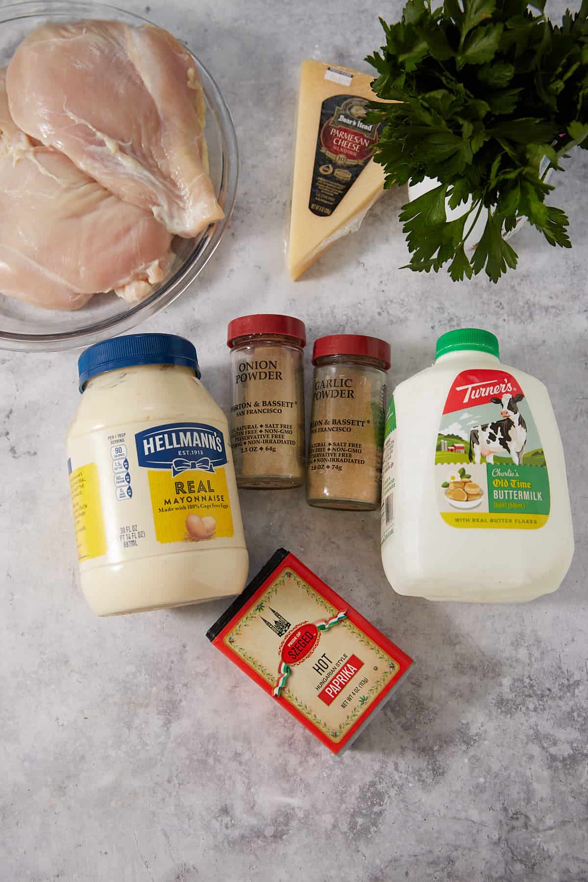 Raw chicken breasts, a wedge of parmesan cheese, fresh parsley, a jar of mayonnaise, jars of onion and garlic powder, a carton of buttermilk, and a box of black pepper arranged on a light gray surface—everything you need for flavor-packed meals or as delicious accompaniments to crowd-pleasing French dip sliders.
