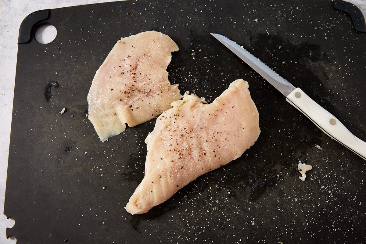 Two raw chicken breasts with salt and pepper on a black cutting board, next to a knife with a white handle—perfectly seasoned and ready for your next recipe, whether it's grilled classics or creative dishes like French dip sliders.