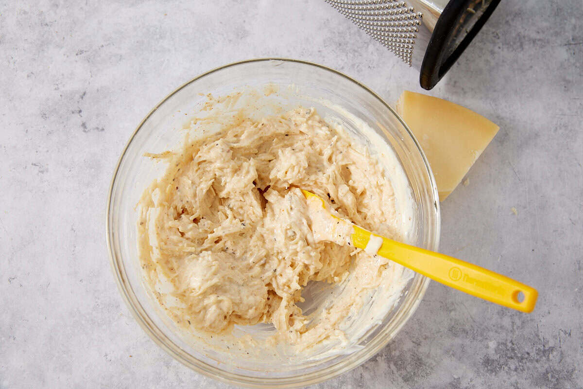 A glass bowl with a creamy, grated mixture and a yellow spatula sits ready to help you assemble your French dip sliders. Nearby, a wedge of cheese and a metal grater rest on a light countertop, setting the stage for this delicious creation.
