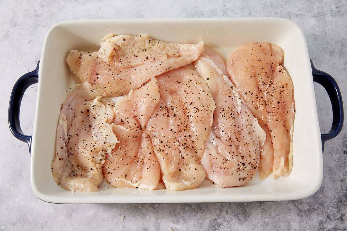 Raw, seasoned chicken breasts, perfect for making French dip sliders, are arranged in a white baking dish with black handles, ready to be cooked. The chicken is sprinkled with black pepper. The background is a light, textured surface.