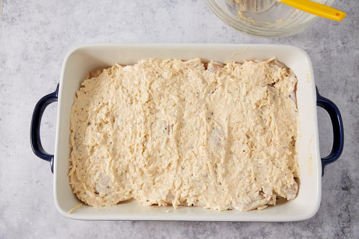 A white baking dish with black handles filled with uncooked chicken covered in a creamy, shredded cheese mixture perfect for making French dip sliders. A glass mixing bowl with a yellow spatula is in the top right corner on a light grey surface.
