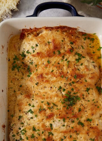 A baked chicken dish in a white casserole dish, topped with melted cheese, browned edges, and sprinkled with chopped parsley—reminiscent of the gooey perfection found in French dip sliders. Nearby are a bowl of grated cheese and some fresh parsley.