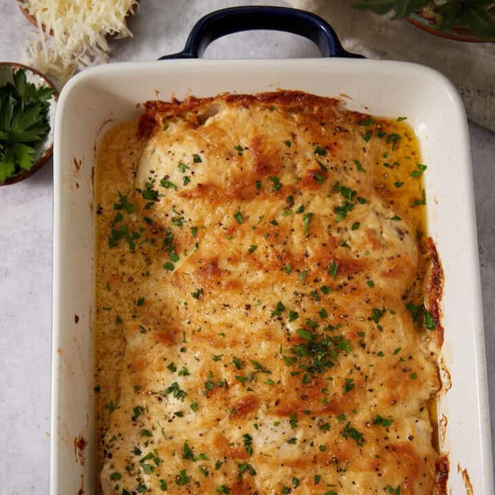 A baked chicken dish in a white casserole dish, topped with melted cheese, browned edges, and sprinkled with chopped parsley—reminiscent of the gooey perfection found in French dip sliders. Nearby are a bowl of grated cheese and some fresh parsley.