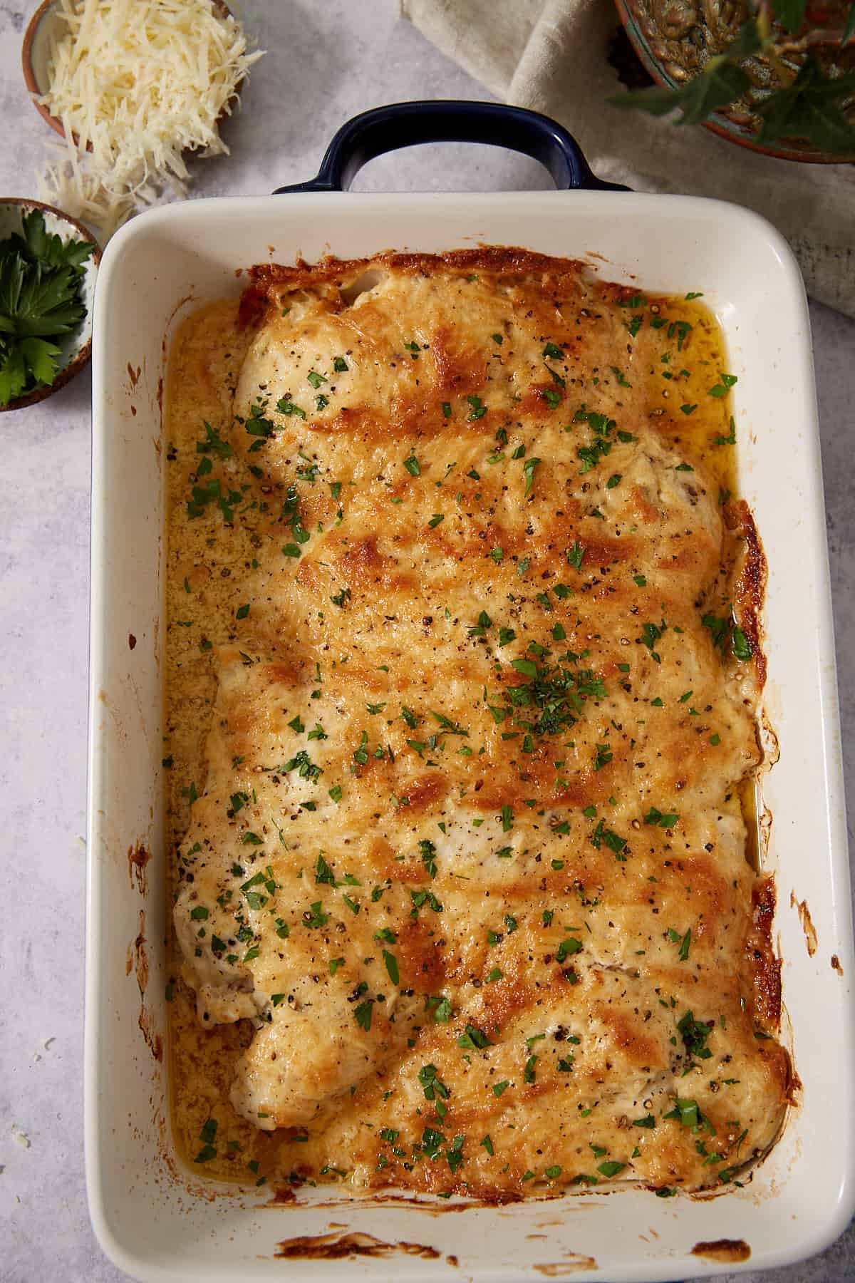 A baked dish of golden, cheesy chicken reminiscent of French dip sliders, topped with fresh chopped parsley in a white rectangular baking pan, with grated cheese and parsley on the side.
