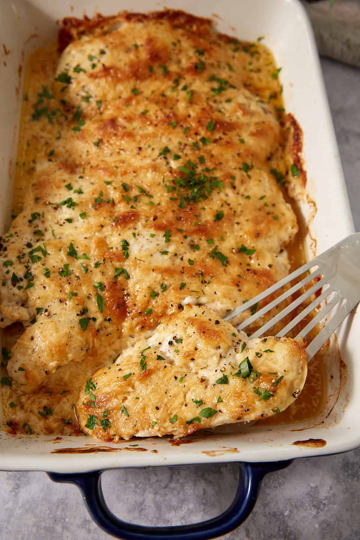Baked chicken breasts topped with herbs and a creamy sauce in a white baking dish, reminiscent of the comforting flavors you love in French dip sliders, with a spatula lifting one piece.