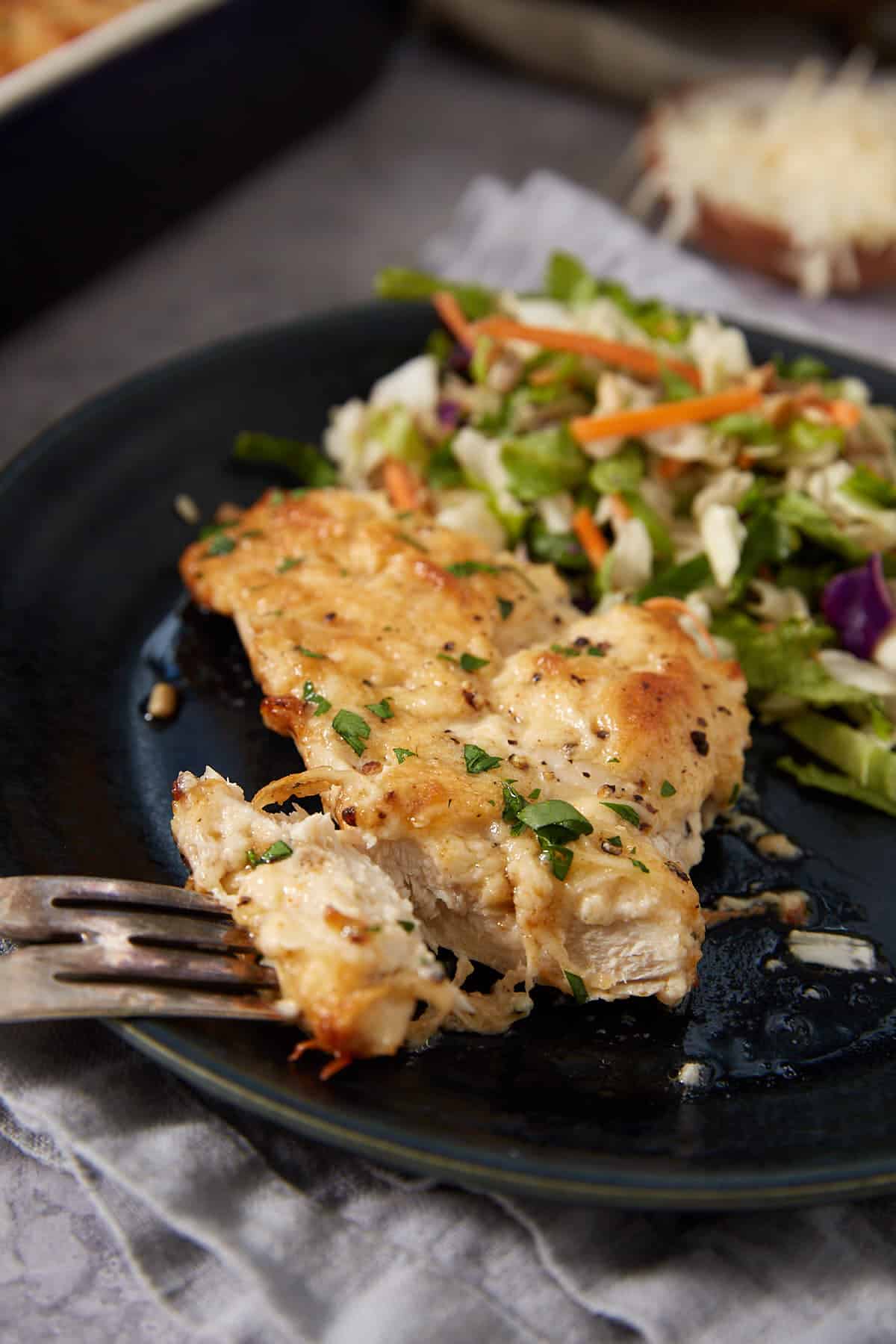 A fork cuts into a piece of golden, baked chicken breast garnished with herbs, served with a fresh vegetable salad on a dark plate, reminiscent of the savory comfort found in classic French dip sliders.