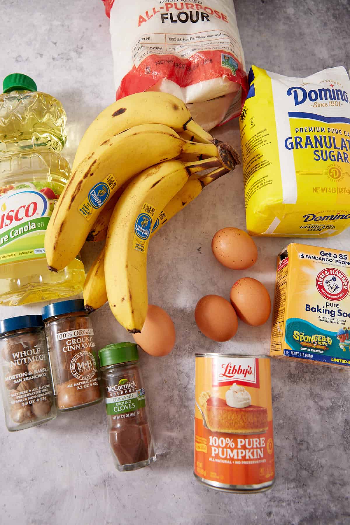 A bunch of bananas, four eggs, a can of pumpkin puree, vegetable oil, flour, sugar, ground cinnamon, ground cloves, baking soda, and baking powder arranged on a gray surface.