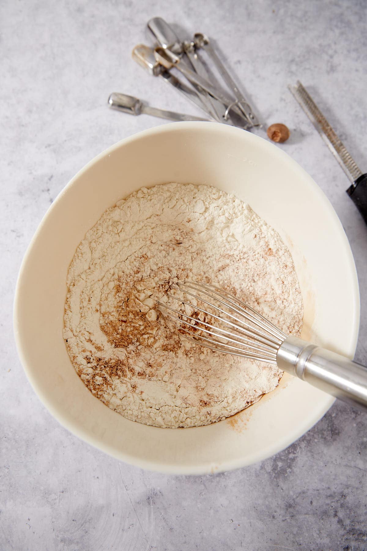 A whisk rests in a white bowl with flour and spices, ready to be mixed. Metal measuring spoons and a grater are on the countertop nearby.