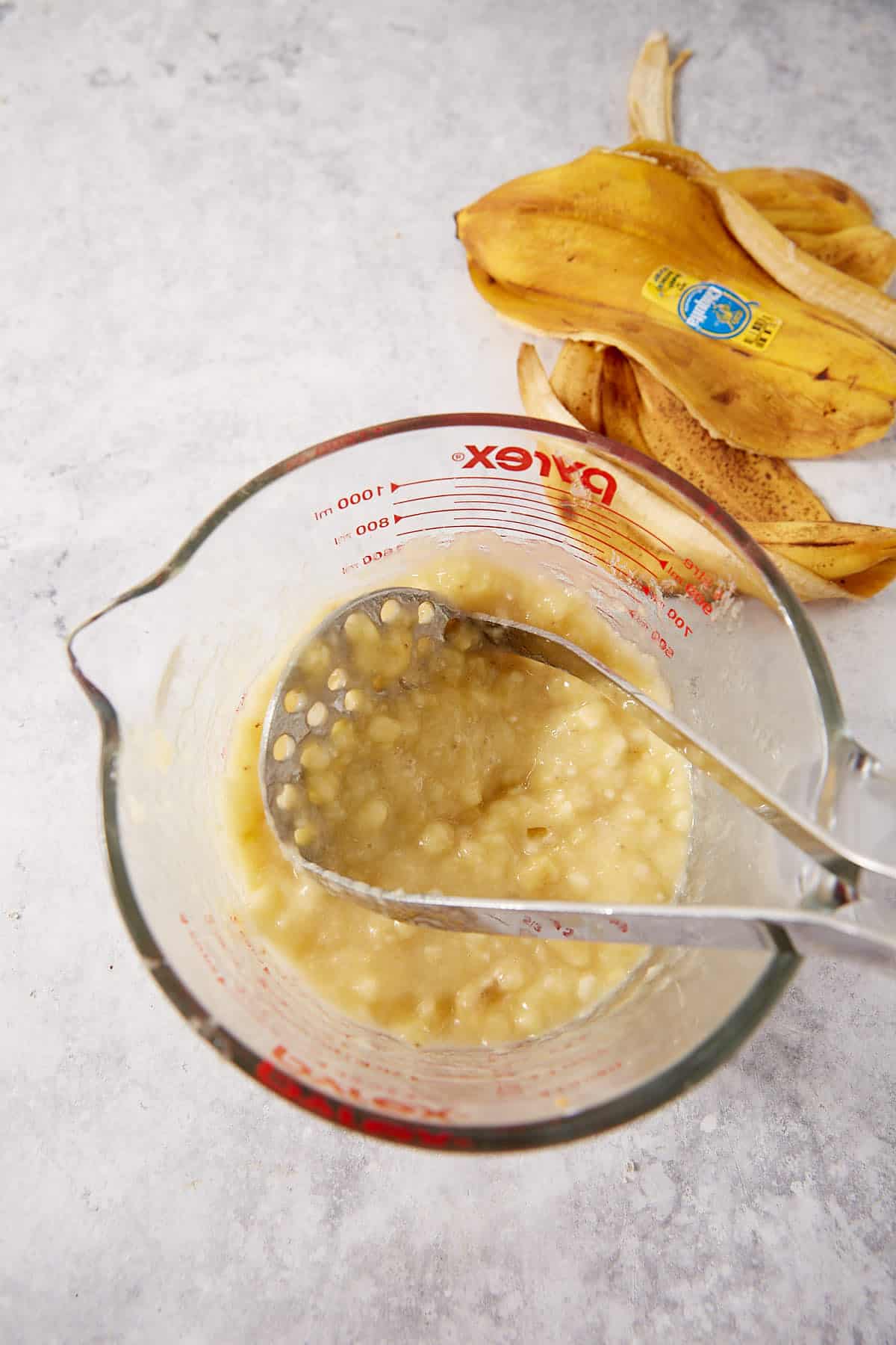 A glass measuring cup with mashed bananas and a metal masher inside, next to a few banana peels on a light surface.