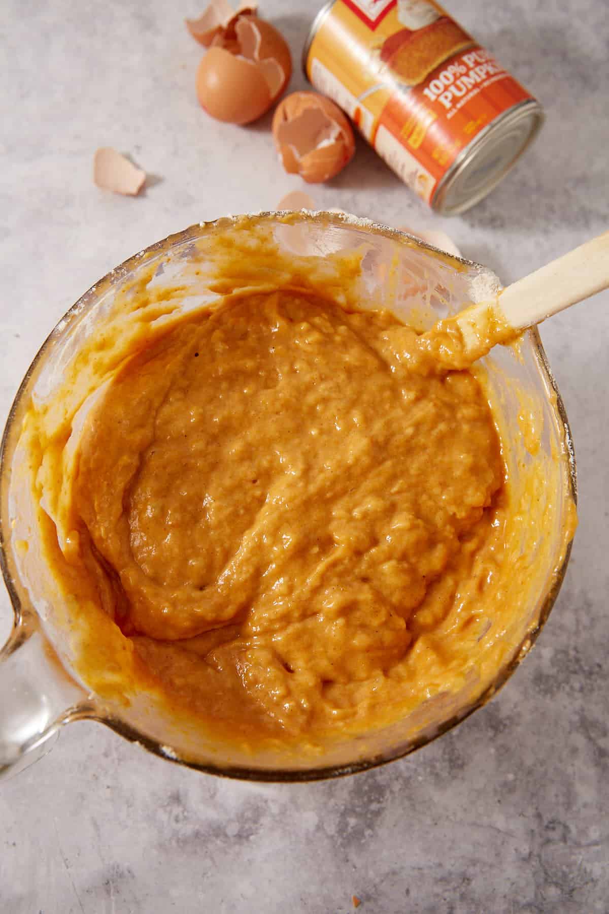 A glass bowl filled with pumpkin batter sits on a counter, with a wooden spoon, eggshells, and a can of pumpkin puree in the background.