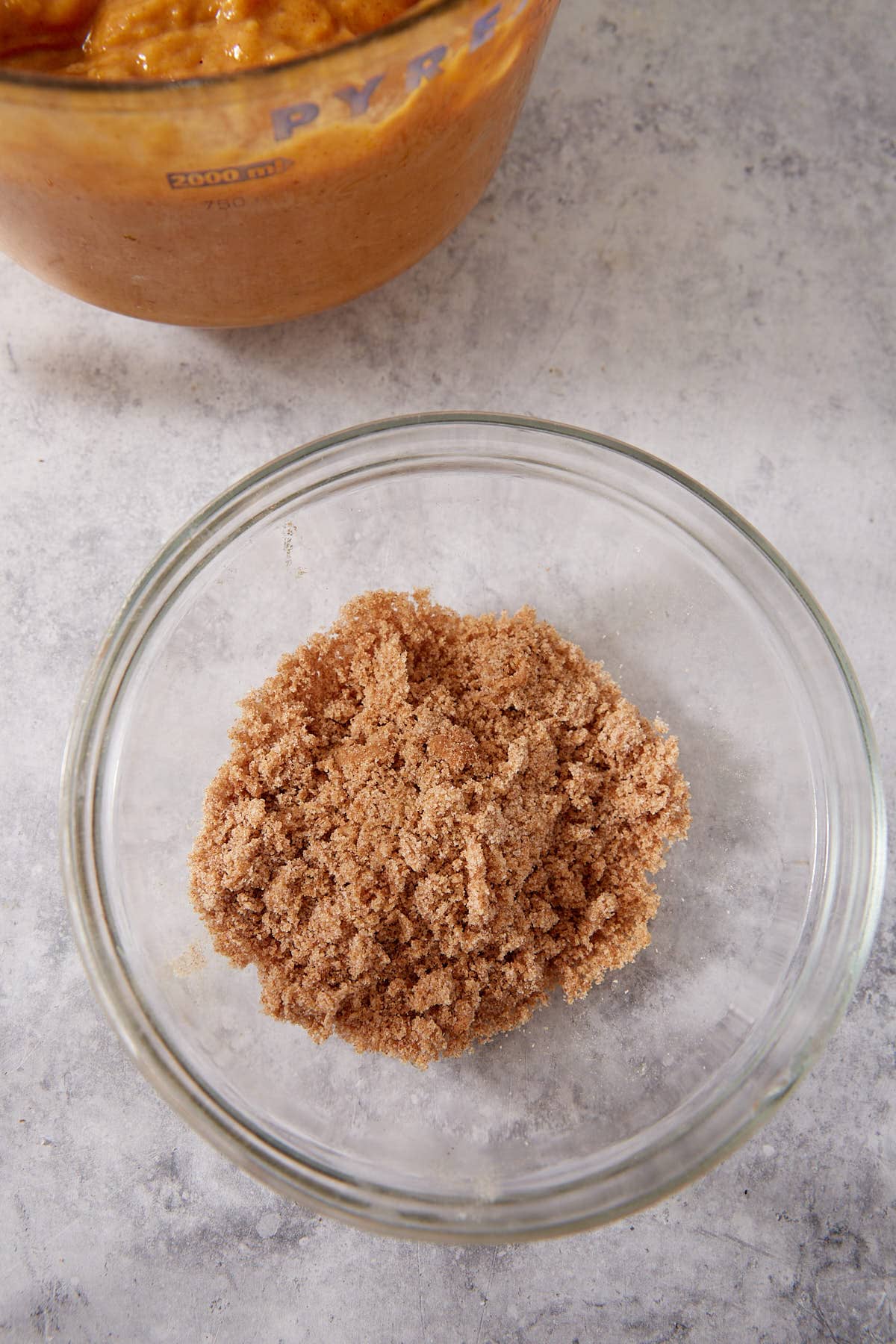A glass bowl filled with a mixture of brown sugar and cinnamon sits on a gray surface. In the background, there is a partial view of a measuring cup with a brown batter inside.