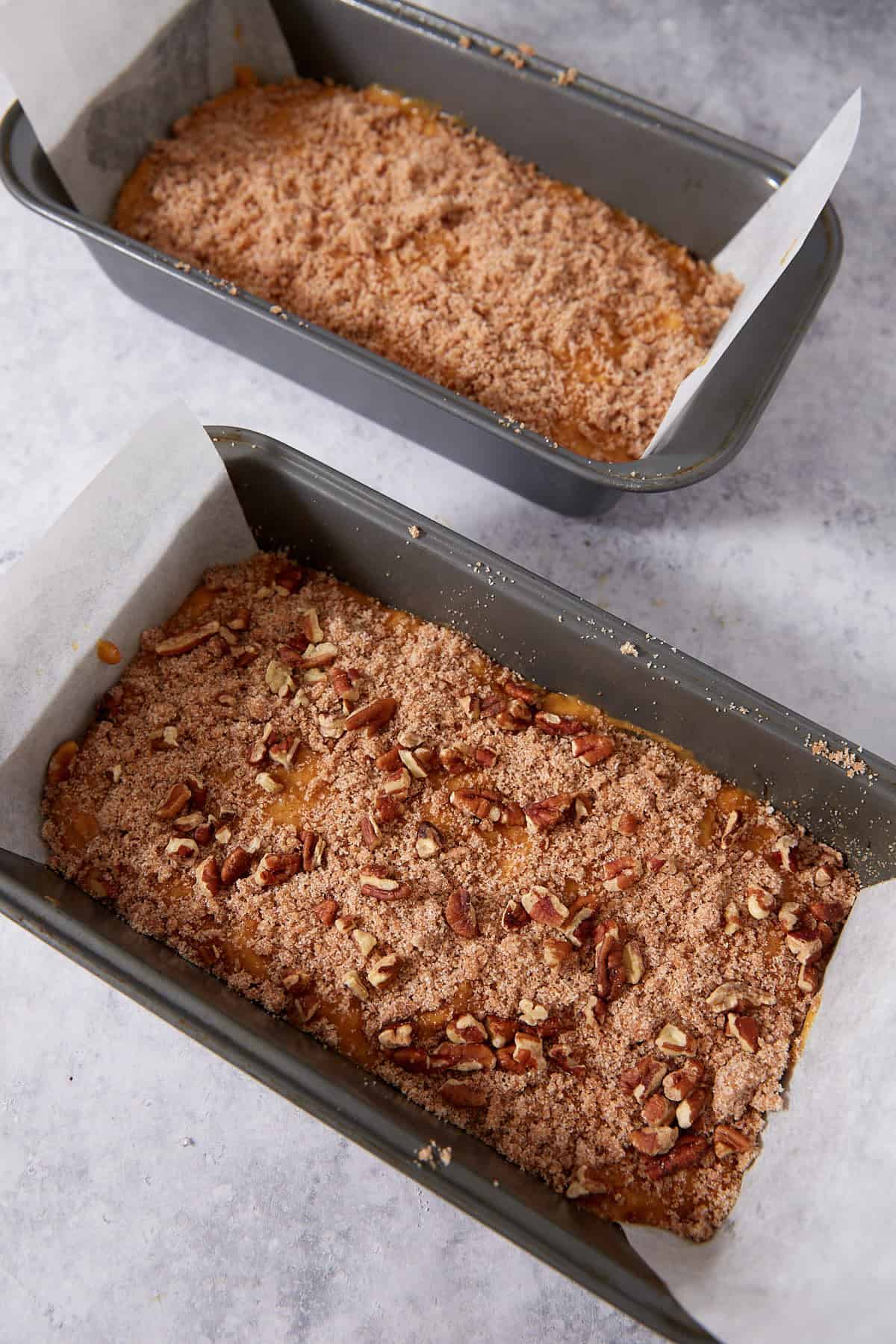 Two loaf pans lined with parchment paper, filled with unbaked batter topped with brown sugar and chopped pecans, ready to be baked. The pans are placed on a light gray countertop.